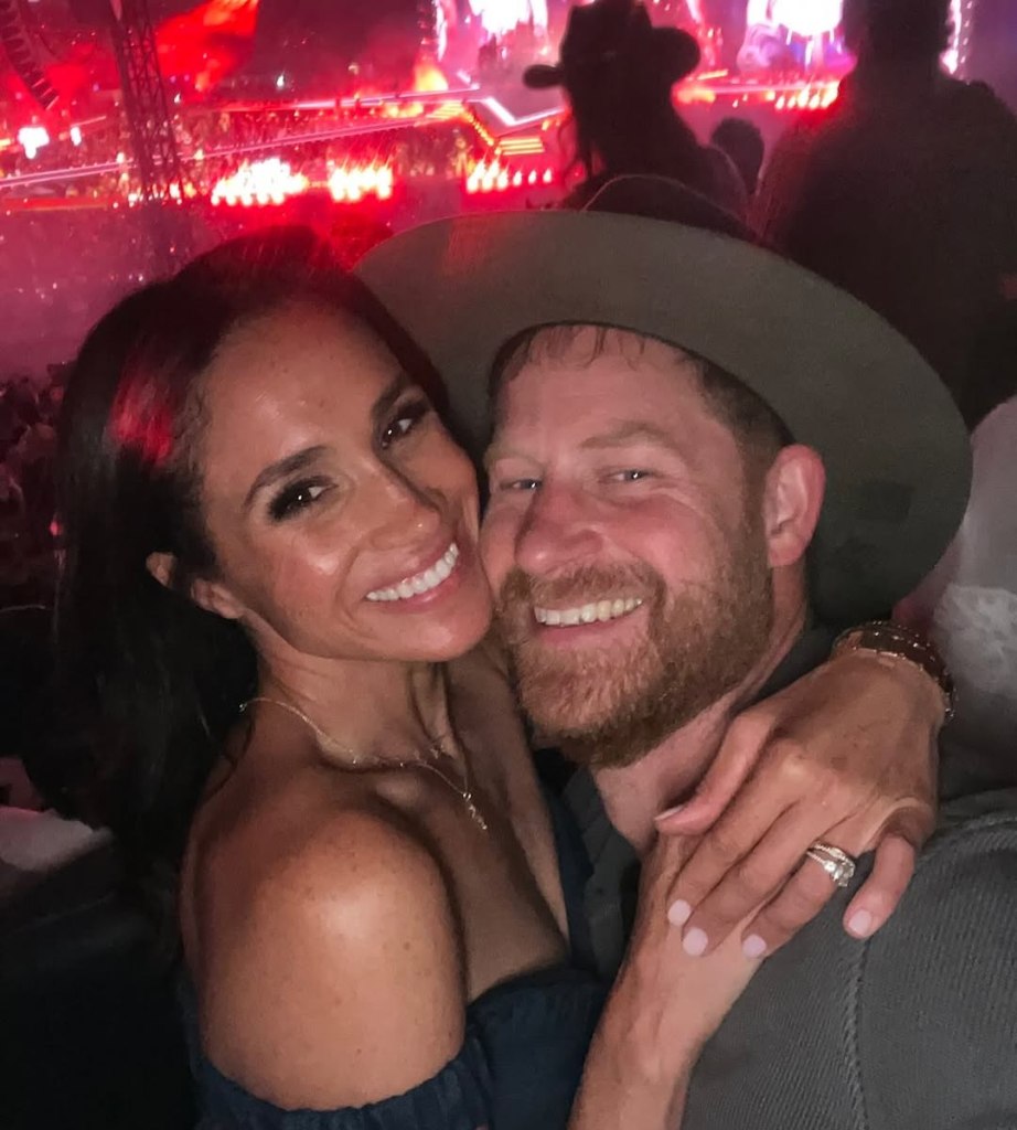 Meghan Markle and Prince Harry smiling and embracing in a crowd with red stage lights in the background.