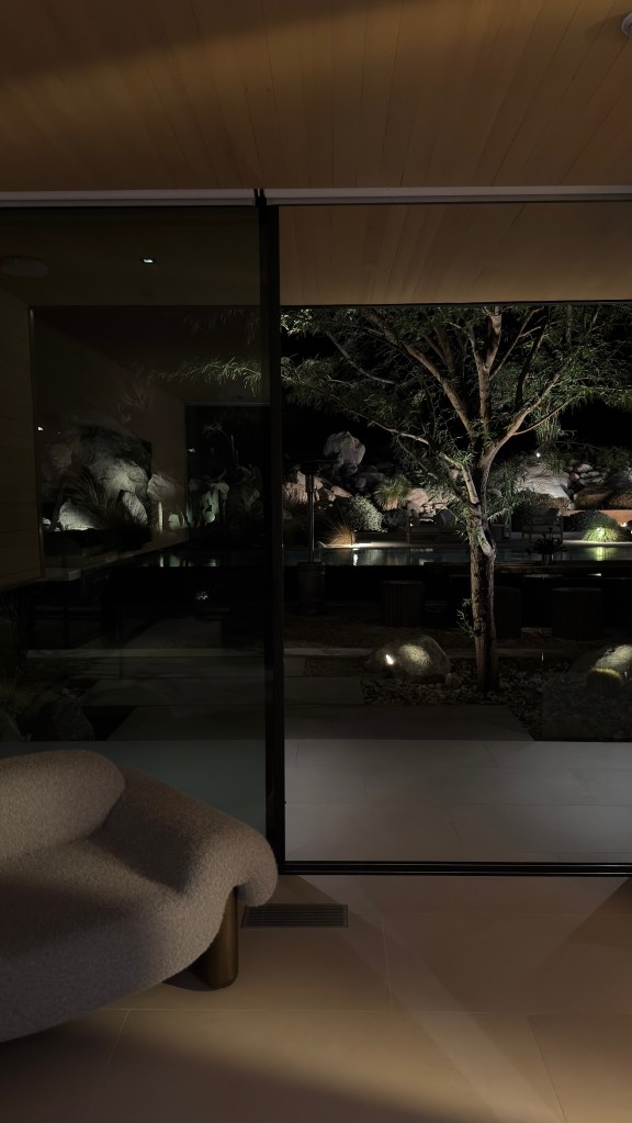 A modern room with a plush white chair in the foreground, looking out to an illuminated outdoor patio at night.