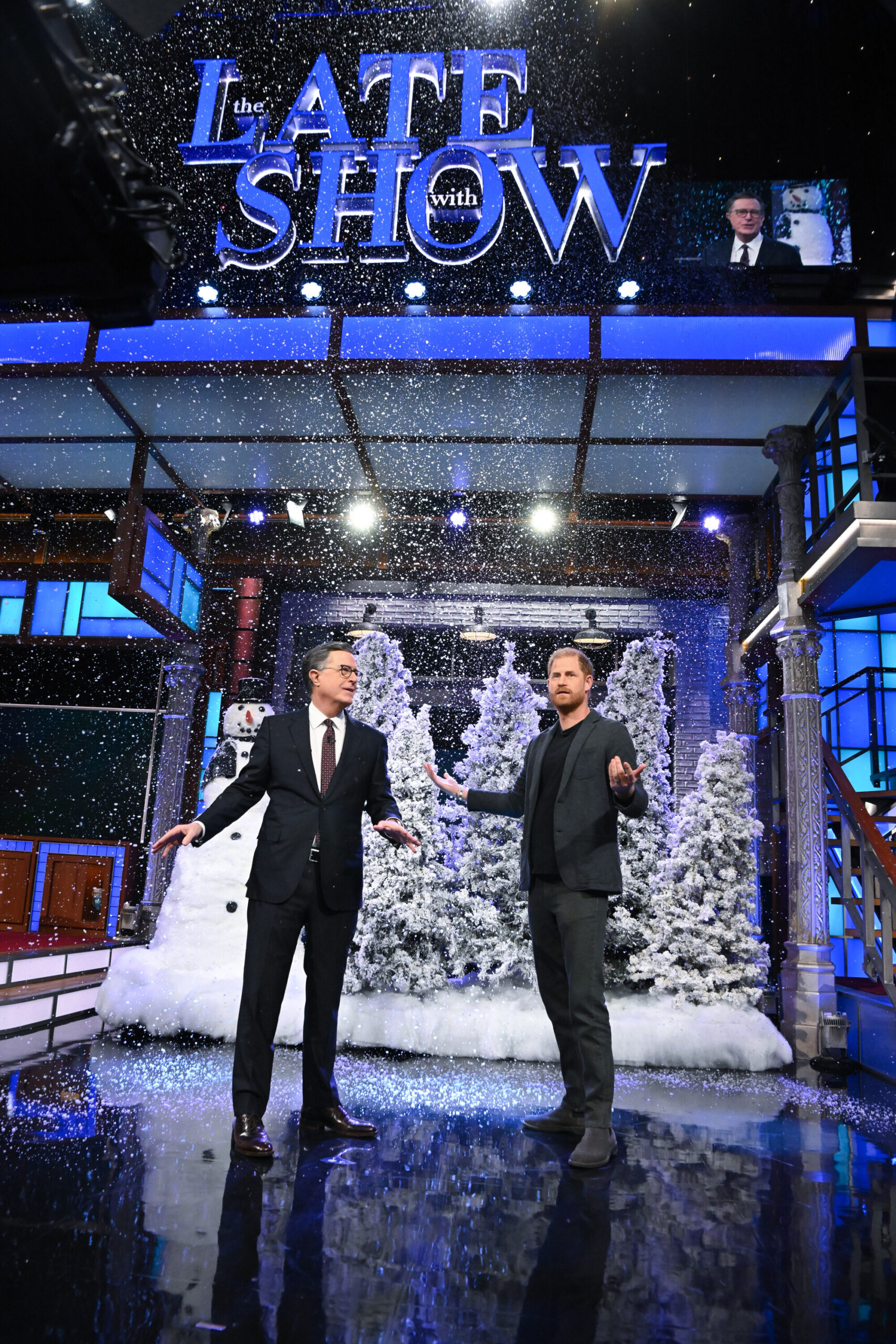 Stephen Colbert and Prince Harry stand on a stage with fake snow falling, surrounded by snowy evergreen trees and a snowman.