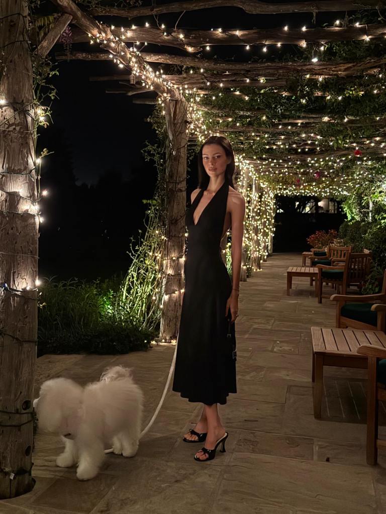 Nicola Peltz in a plunging black dress on a patio at night beside a white dog.