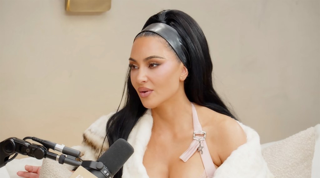 Kim Kardashian, with a black ponytail and a black headband, sitting in a beige setting, wearing a pink bra and a fur coat.