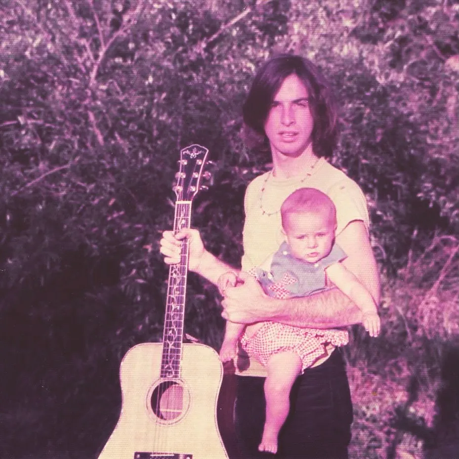 Robert Carradine holding baby Ever while resting an acoustic guitar on his lap.