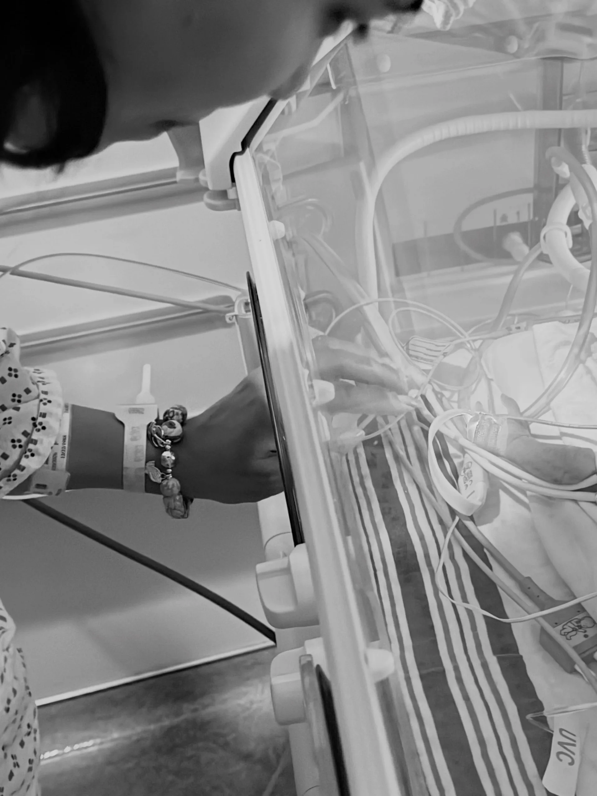A parent's hand reaches into a NICU incubator to touch newborn Aidan, with monitoring wires visible.