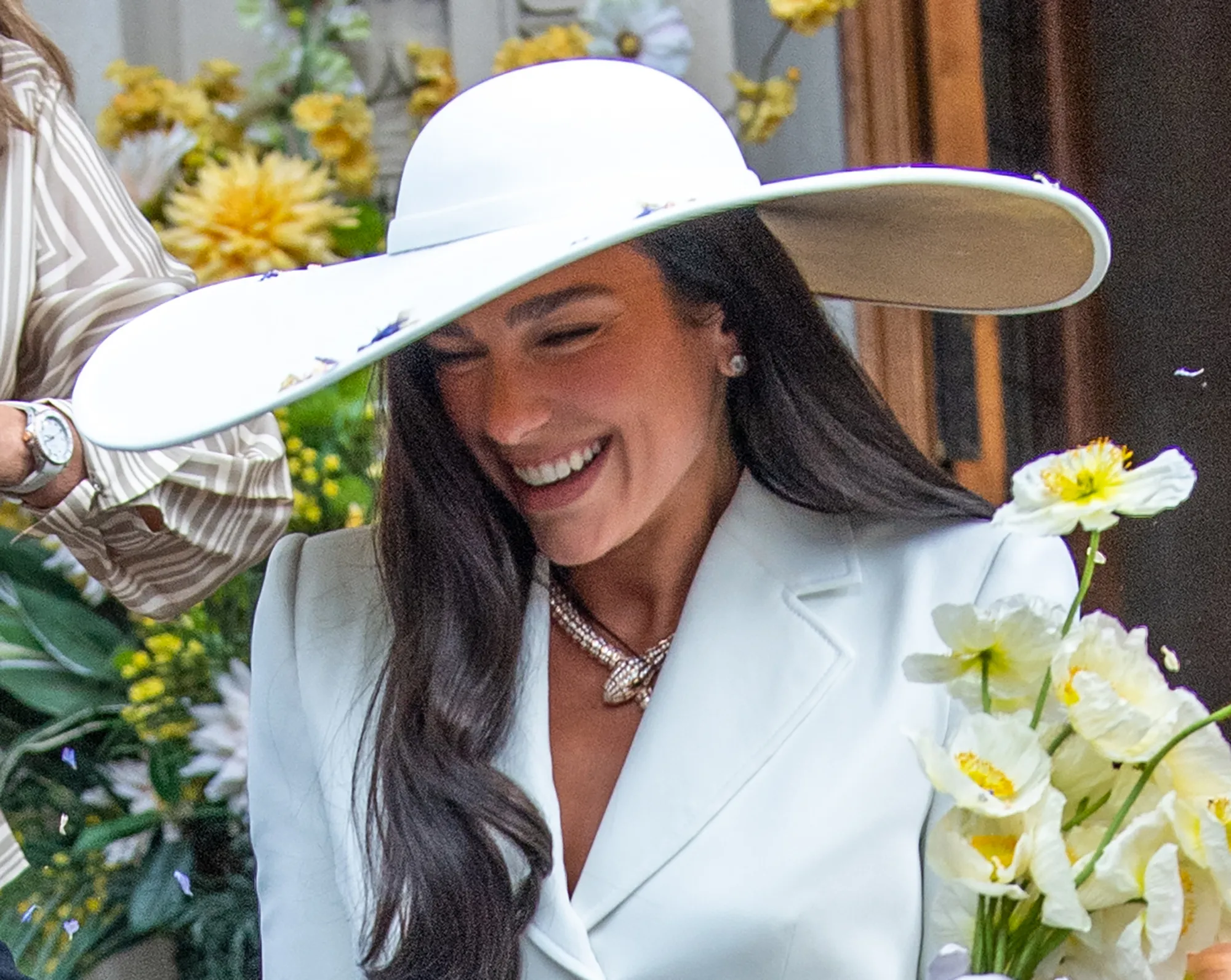 Dua Lipa in an ivory Schiaparelli suit and wide-brim hat, wearing a Bulgari Serpenti necklace and holding white flowers.
